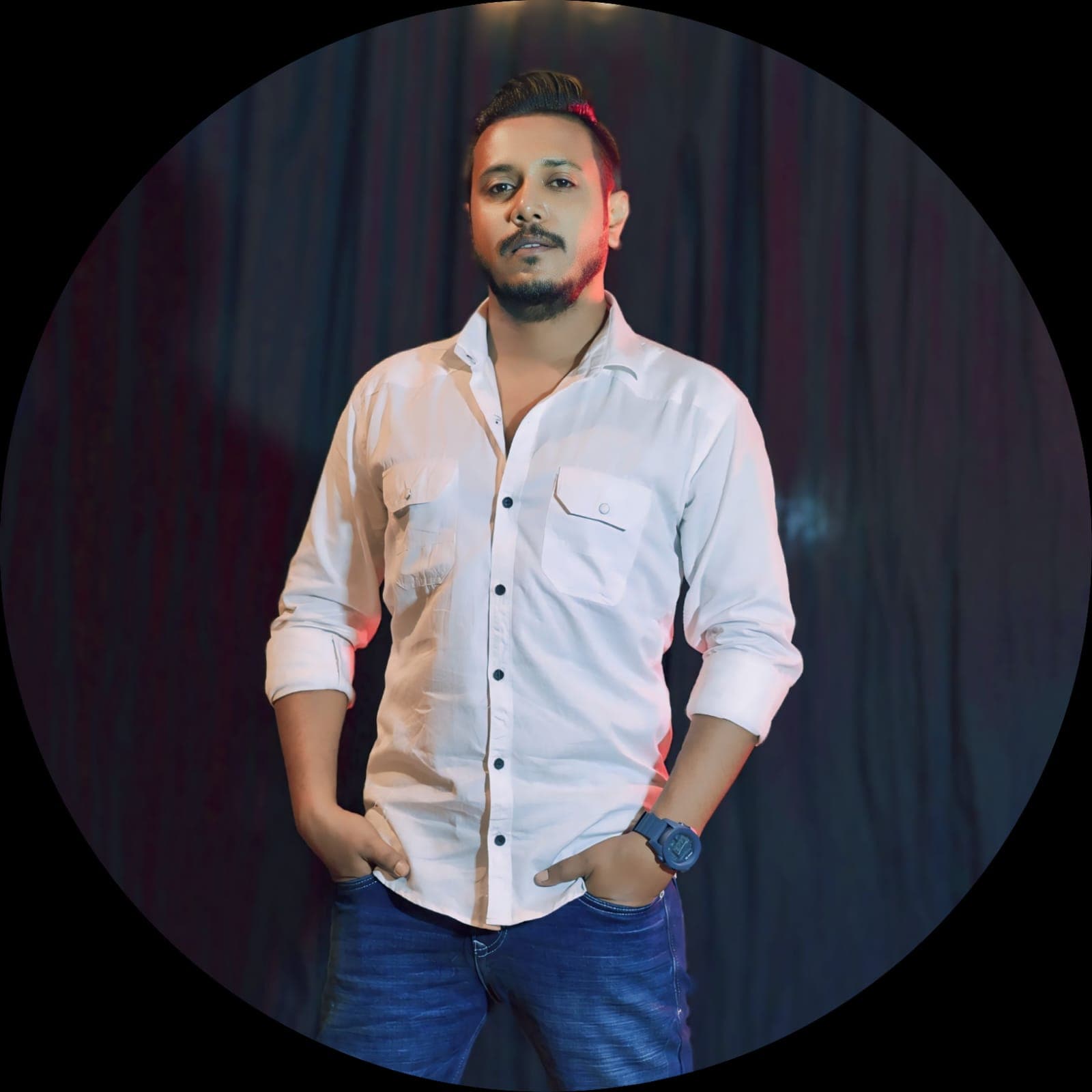 Confident man with beard, white shirt, blue jeans, hands in pockets, dark background.